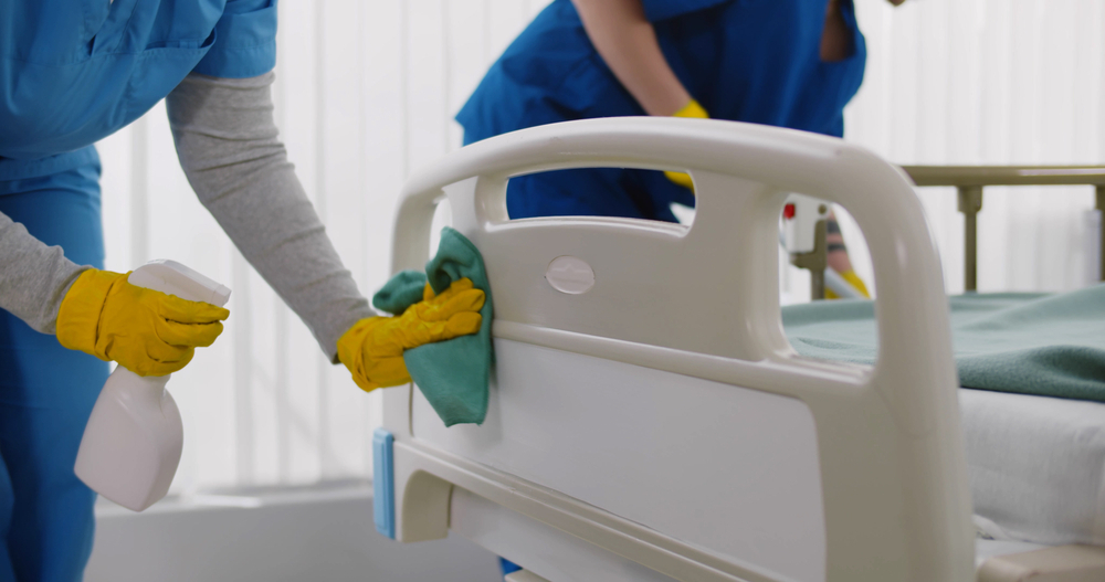 Zwei Reinigungskräfte desinfizieren ein Krankenbett in einer Praxis mit Sprühflasche und Tuch, während gelbe Handschuhe und blaue Kleidung auffallen.