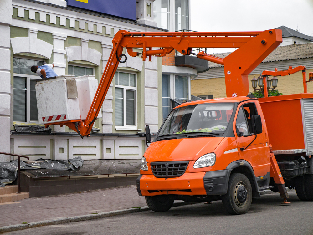 FassadenNEW Orangefarbenes Fahrzeug mit hoher Arbeitsbühne reinigt die Fassade eines mehrstöckigen Gebäudes in der Innenstadt.