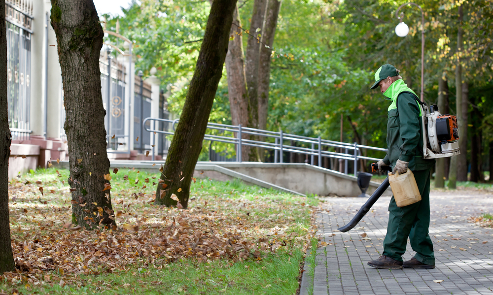 Hausmeisternew Mitarbeiter im grünen Arbeitsanzug bläst Laub auf einem Gehweg neben einem gepflegten Gebäude weg.